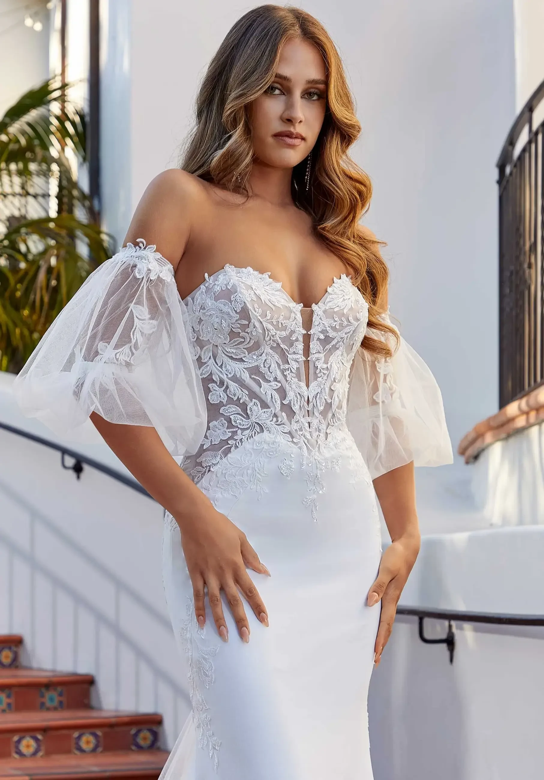 Model is wearing a strapless fitted wedding dress with detachable puff sleeves and intricate lace on the bodice