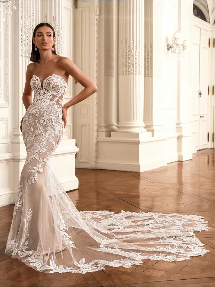 A woman in an elegant, strapless, lace wedding gown with a long train stands in a grand hall with ornate white pillars and wood flooring, embodying grace.