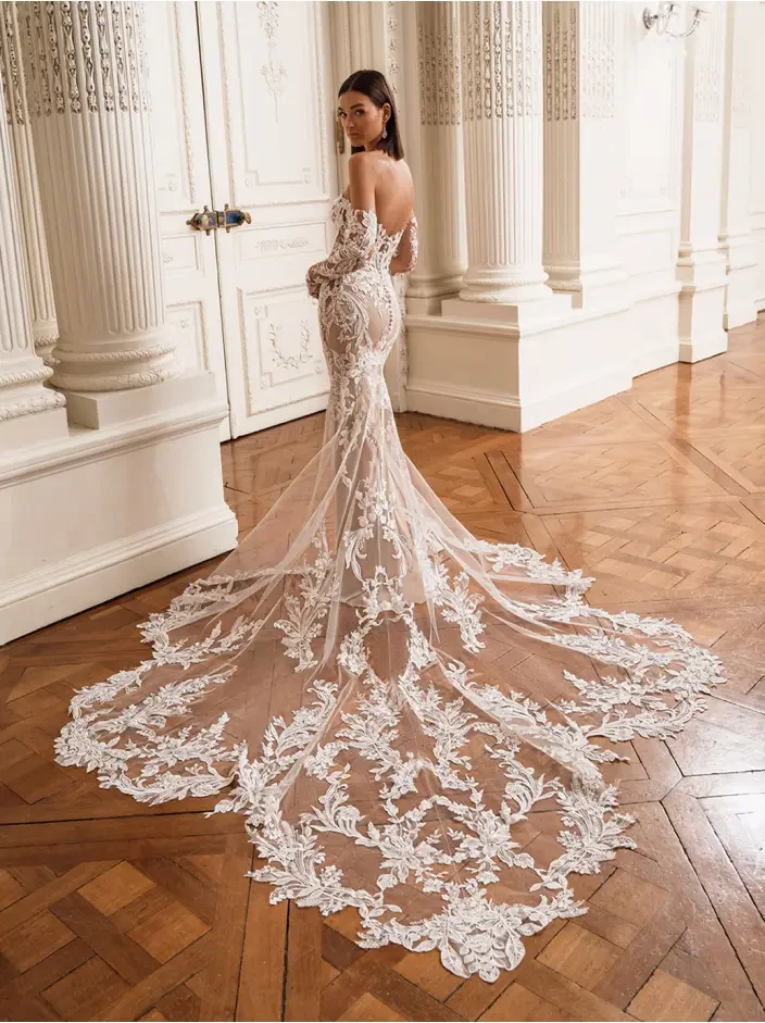 A bride in a lace wedding gown with a long, embroidered train stands in an ornate room with wooden floors and white columns, exuding elegance and grace.