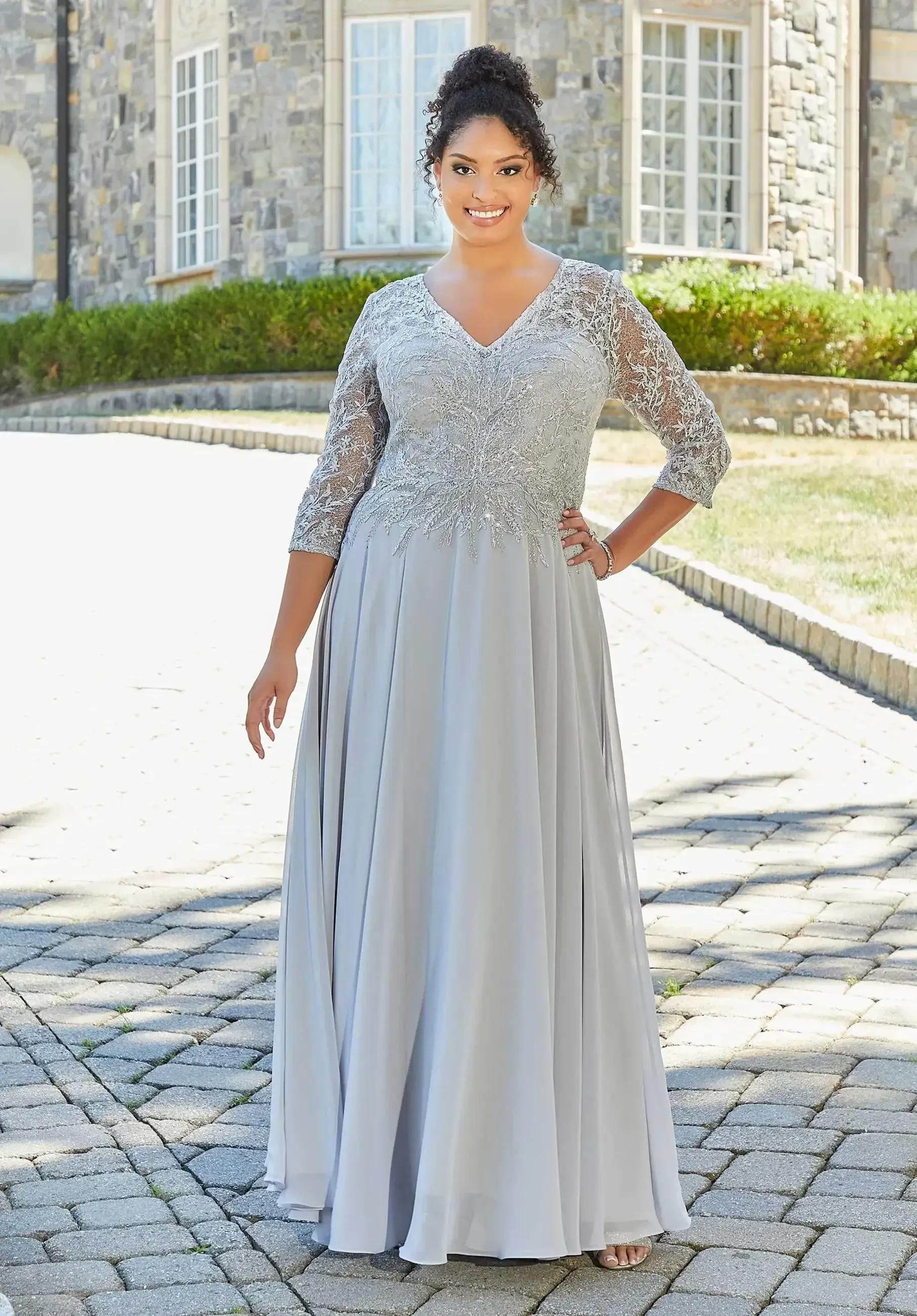 A woman wearing a long, silver-grey dress with lace sleeves, smiling and posing outdoors on a stone pathway.