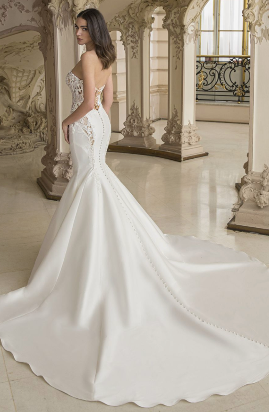 A woman in an elegant white mermaid wedding dress with lace details and a long train stands in an ornate, classic room, exuding a sophisticated, serene vibe.