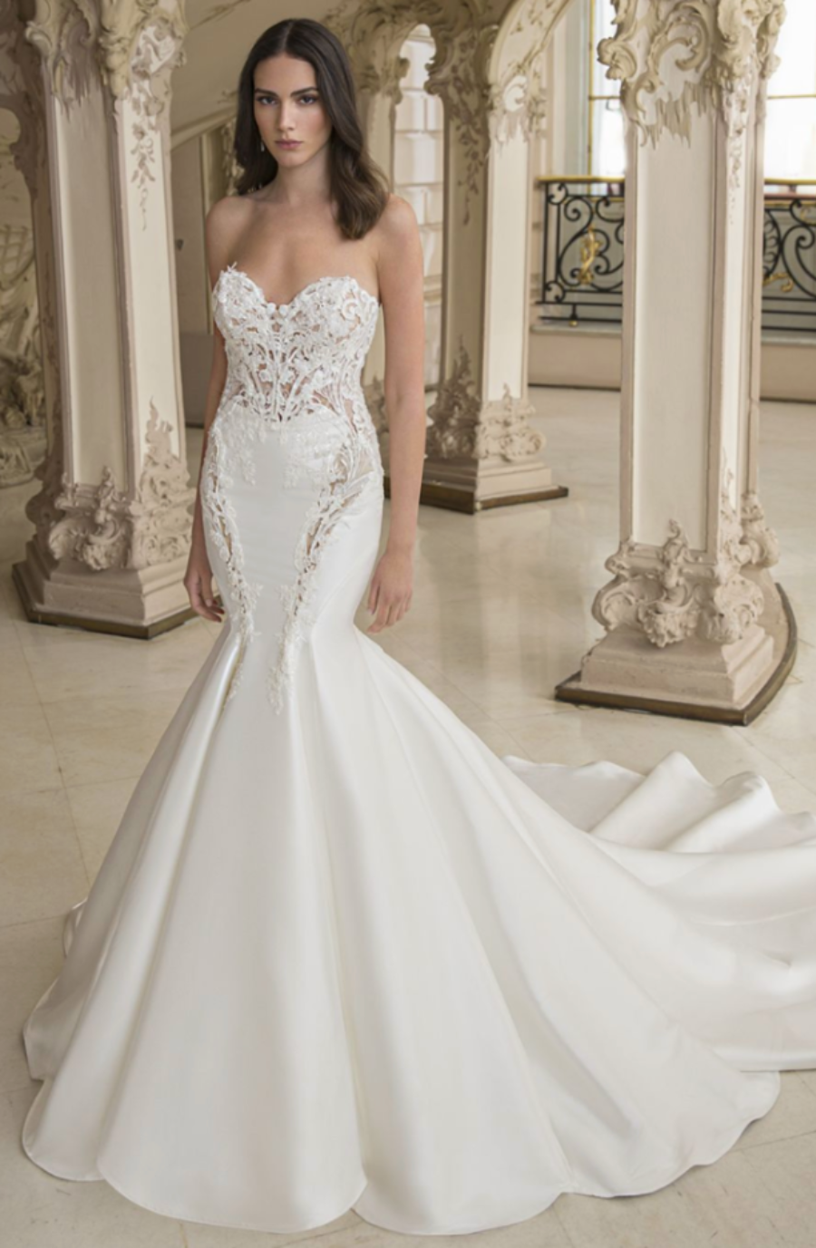 A woman in an elegant, strapless white mermaid gown with lace detailing poses in an ornate, vintage-style room with intricate arches, exuding grace and sophistication.