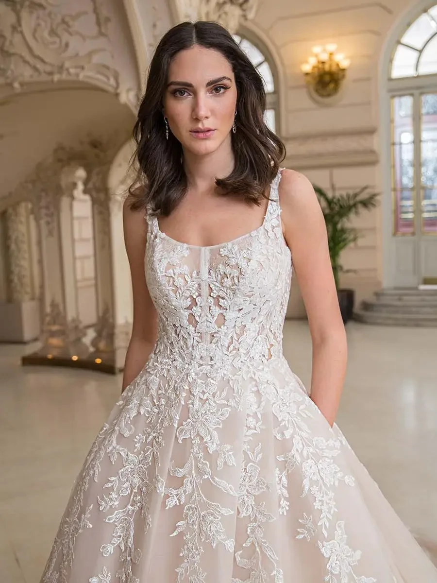 A woman in an ornate wedding gown stands in an elegant room with arched decor and soft lighting. Her dress features intricate floral lace.