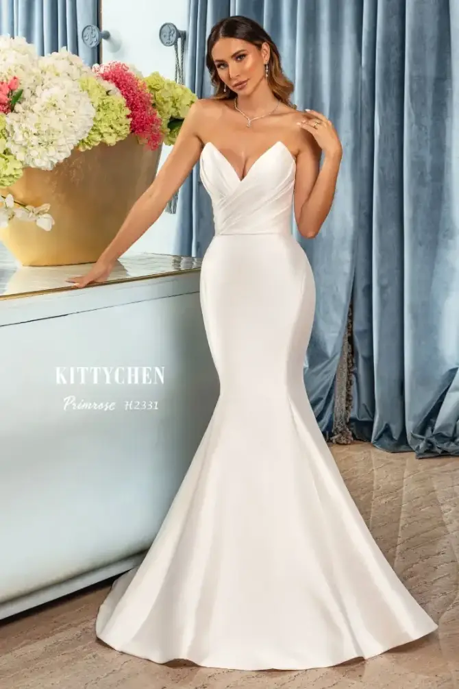 A woman in a fitted white gown with a sweetheart neckline stands beside a large vase of flowers, in front of a light blue backdrop. The dress has an elegant mermaid silhouette.