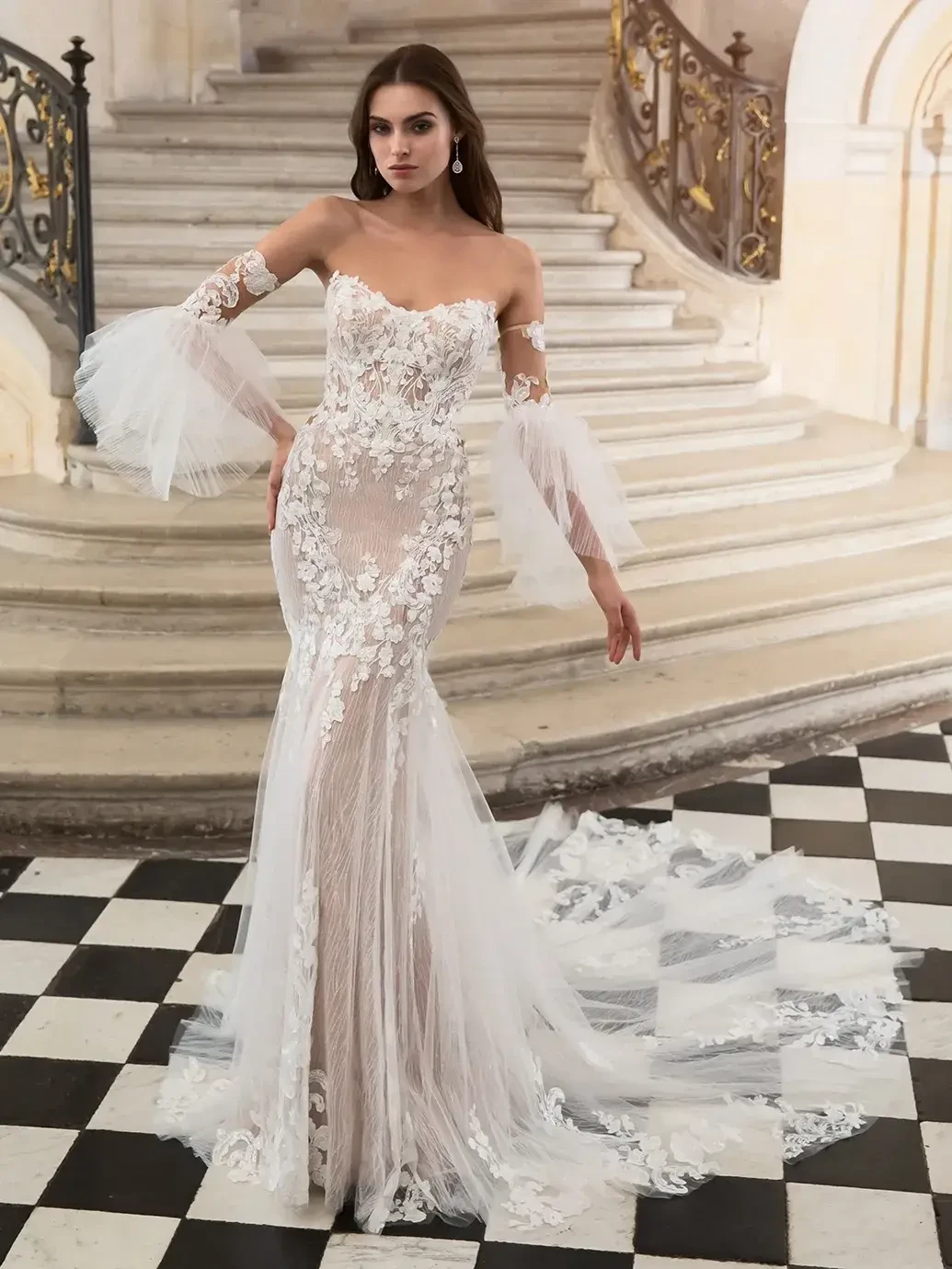 A model wears a long, elegant, off-shoulder wedding dress adorned with floral details and sheer sleeves, posing on a checkered floor in an ornate staircase setting.