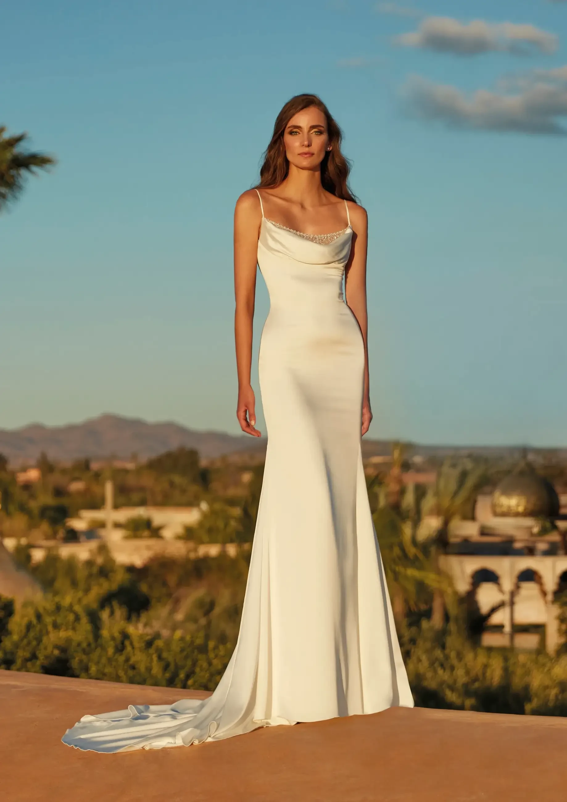 Elegant woman in a long, fitted white gown with spaghetti straps, standing against a serene, sunny desert backdrop with distant mountains and palm trees.