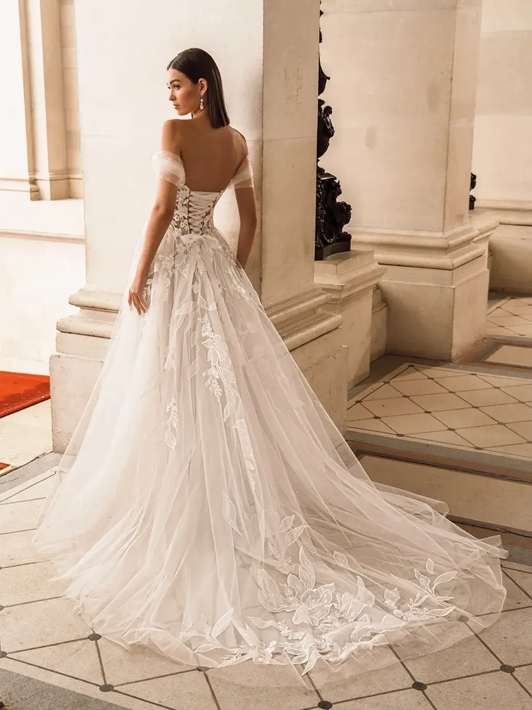 A woman stands gracefully in an elegant, lace-embellished wedding gown with a flowing train, set against a grand, classical architectural backdrop.