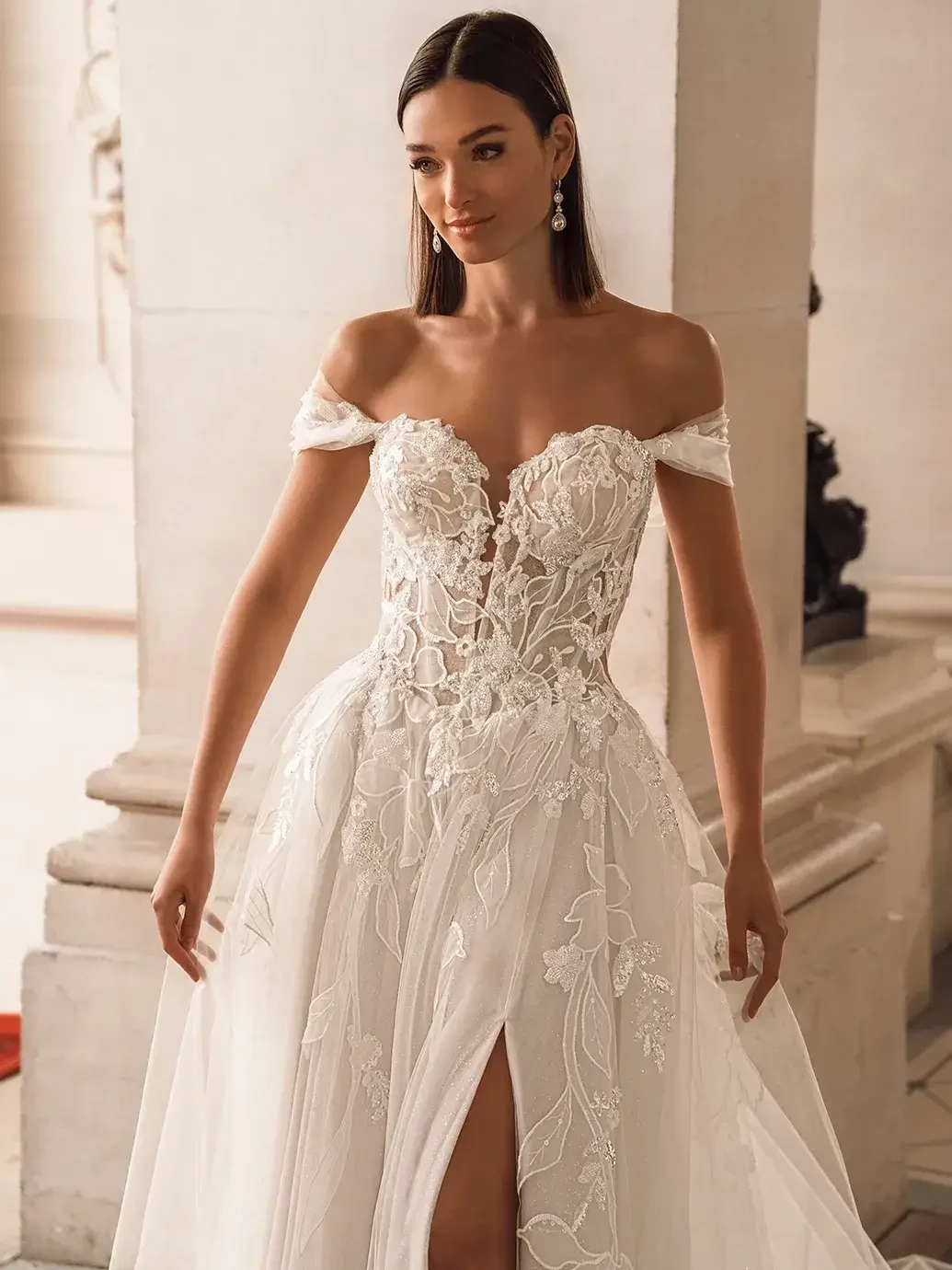 A woman in an elegant off-the-shoulder wedding gown with floral lace details and a thigh-high slit stands in a bright, stately room, exuding a serene and joyful aura.