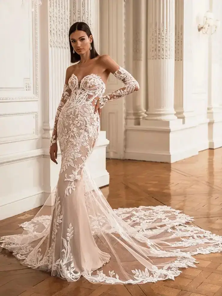 Bridal gown with intricate lace, sheer sleeves, and an elegant train in a grand room with white columns and parquet floor, exuding sophistication.