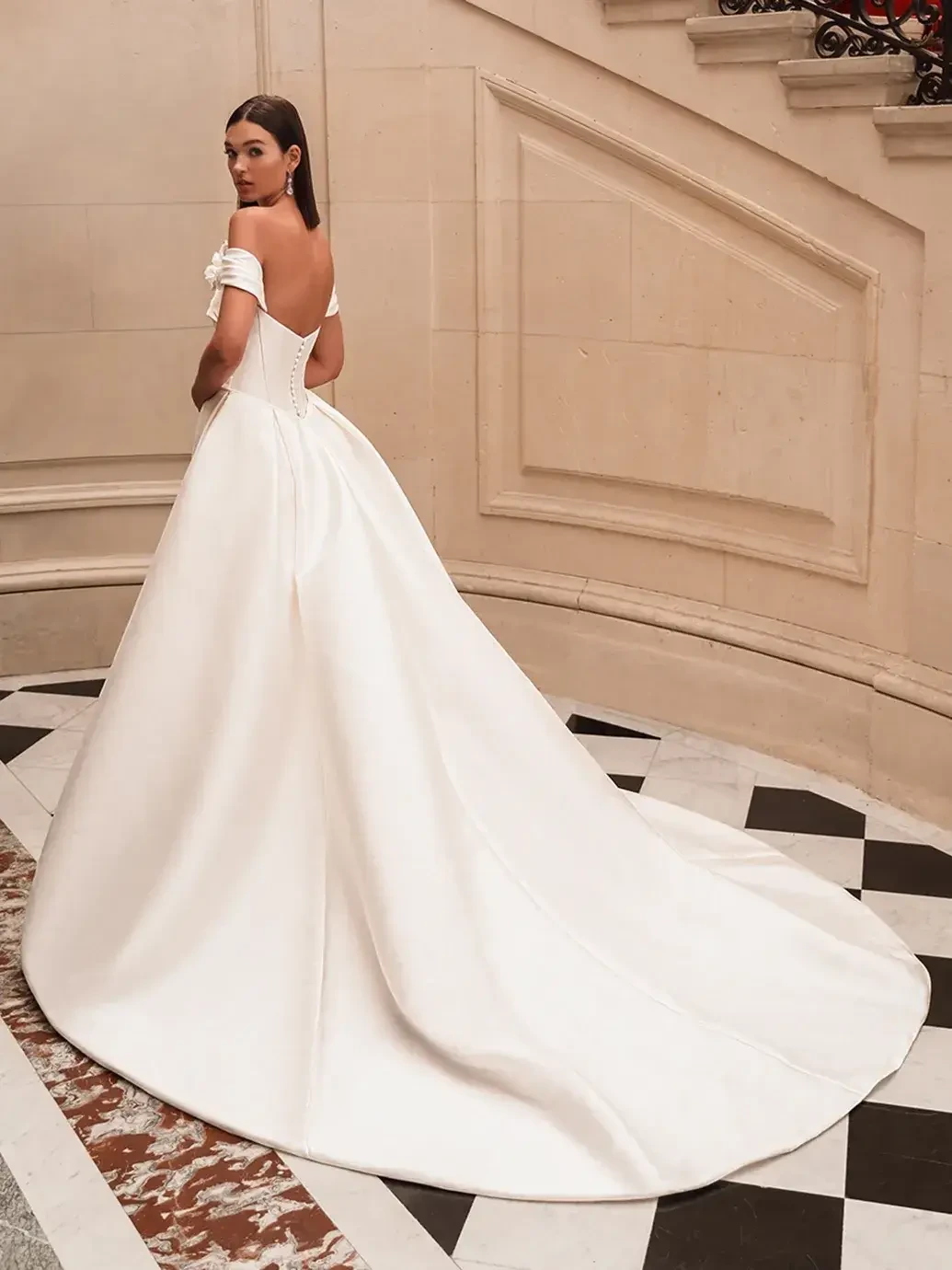 A woman in an elegant, white off-shoulder gown stands on a checkered marble floor by a grand staircase. The long train adds a touch of luxury and timeless elegance.